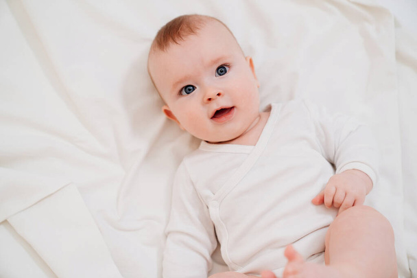 vauva valkoisissa vaatteissa makaa selällään sängyllä
 - Valokuva, kuva