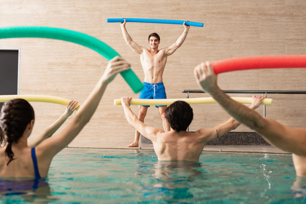 Selective focus of handsome trainer smiling while training group of young people with pool noodles in swimming pool - Photo, Image