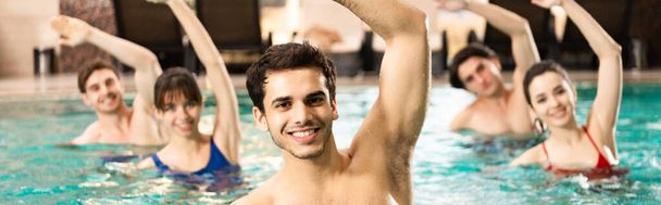 Selective focus of handsome trainer smiling at camera while exercising with people in swimming pool, panoramic shot - Photo, Image