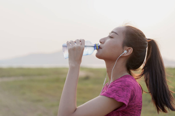 Sivunäkymä Aasian kunto urheilija naisten juoda pullon vettä ja rentoutua harjoituksen jälkeen tai käynnissä kesällä auringonlaskun tai auringonnousun. Käsitteen hyvinvointi ja elämäntapa
 - Valokuva, kuva