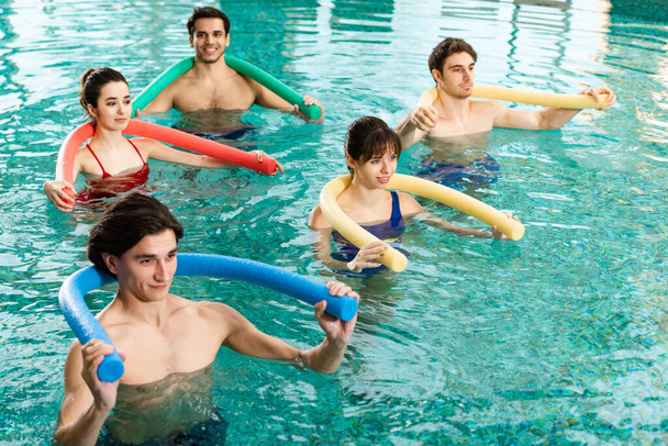 Smiling young people exercising with pool noodles during water aerobics in swimming pool - Photo, Image