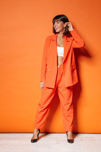 Full length portrait of a beautiful smiling young brown short haired woman wearing suit and high heels standing over orange background, posing - Foto, Imagem