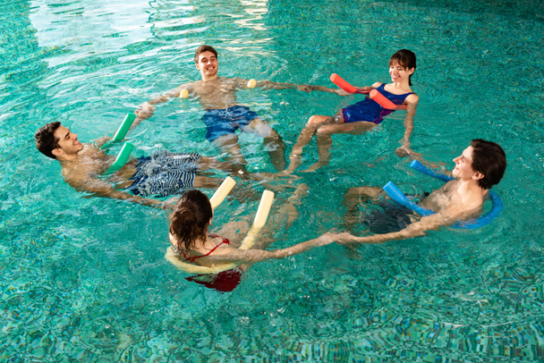 High angle view of smiling young people holding hands while exercising with pool noodles in swimming pool - Photo, Image