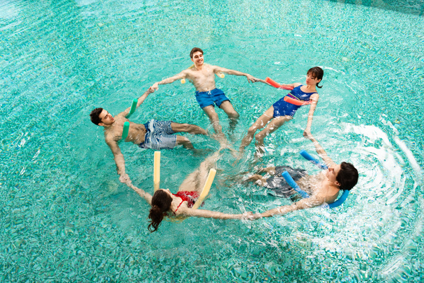 Wysoki kąt widzenia grupy młodych ludzi trzymających się za ręce podczas treningu aerobiku wodnego z makaronem basenowym w basenie - Zdjęcie, obraz