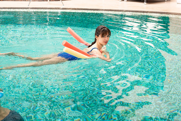 Zijaanzicht van jonge vrouw op zoek naar camera tijdens het zwemmen met zwembad noedels in het zwembad - Foto, afbeelding