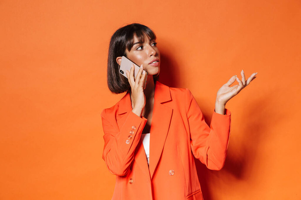 Portrait of a beautiful young brown short haired woman wearing suit standing over orange background, talking on mobile phone, looking away - Фото, изображение