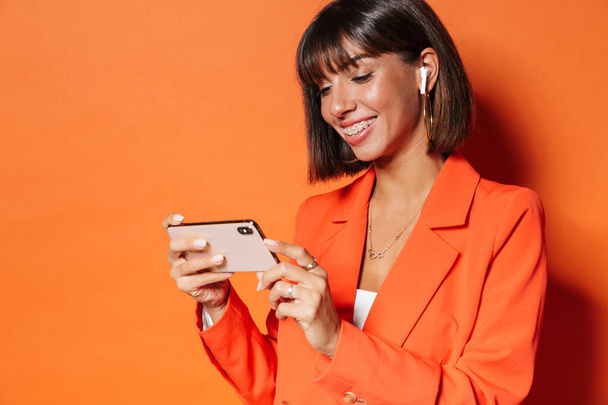Gyönyörű mosolygós fiatal nő visel kabát áll elszigetelt narancssárga háttér, a mobiltelefon, videó megtekintése - Fotó, kép