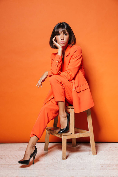 Full length of a beautiful young woman wearing suit sitting on a chair isolated over orange background, posing, looking at camera - Foto, Bild