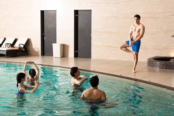 Yüzme havuzunda genç insanlara egzersiz yaparken tek ayak üstünde duran yakışıklı antrenör. - Fotoğraf, Görsel