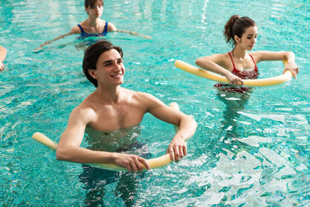 Selective focus of smiling man holding pool noodles while training with women in swimming pool - Foto, imagen