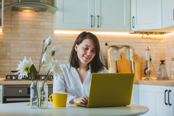 young pretty caucasian woman talking on the phone sitting in front of laptop in the kitchen. business housewife. drinking coffee from yellow mug - Foto, immagini