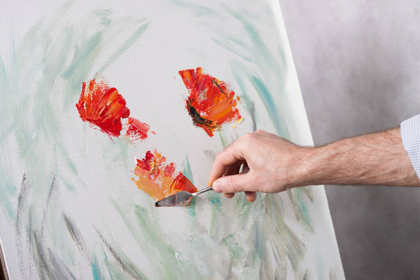An artist with a palette knife paints red flowers on a white canvas. The beginning of a picturesque picture - Fotó, kép