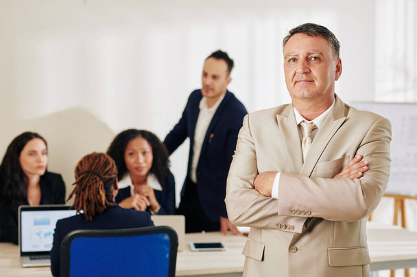Zelfverzekerde succesvolle volwassen zakenman staat met zijn armen gevouwen, zijn medewerkers bespreken project op de achtergrond - Foto, afbeelding