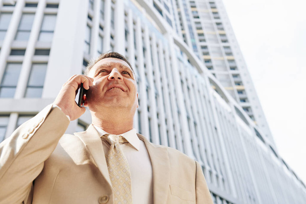 Biznesmen w średnim wieku stojący przed budynkiem biurowym i rozmawiający przez telefon z partnerem biznesowym - Zdjęcie, obraz