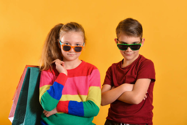 Boy and girl in sun glasses and with shopping bags, on a yellow background. - Foto, immagini