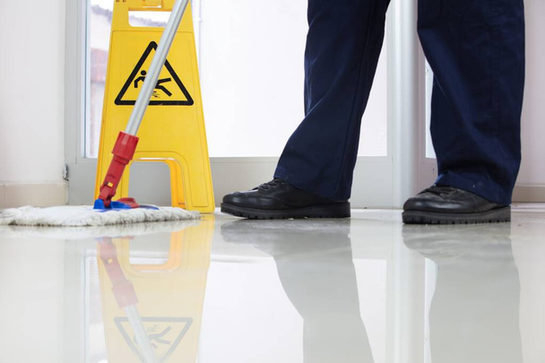 A low angle closeup of a person cleaning the floor with a mop near a yellow caution wet floor sign - Foto, Bild