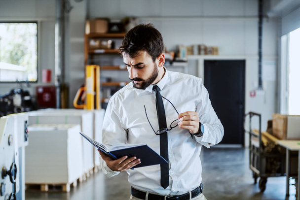 Handsome caucasian serious bearded graphic engineer in shirt and tie, holding notebook and looking at it while standing in printing shop. - Zdjęcie, obraz