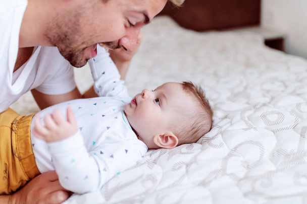 Close up of cute six months old baby boy lying on bed and playing with his dad. Dad talking to son and cuddling him. - Photo, Image