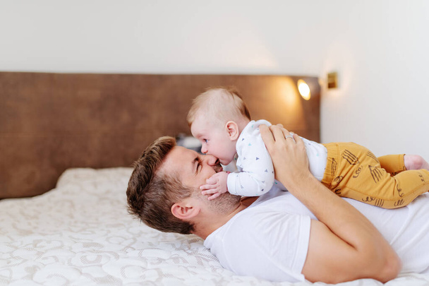 Glimlachende blanke knappe vader liggend op bed in de slaapkamer en met zijn liefdevolle schattige zes maanden oude zoon op zijn borst. De baby bijt op papa 's neus. Onvoorwaardelijke liefde concept. - Foto, afbeelding