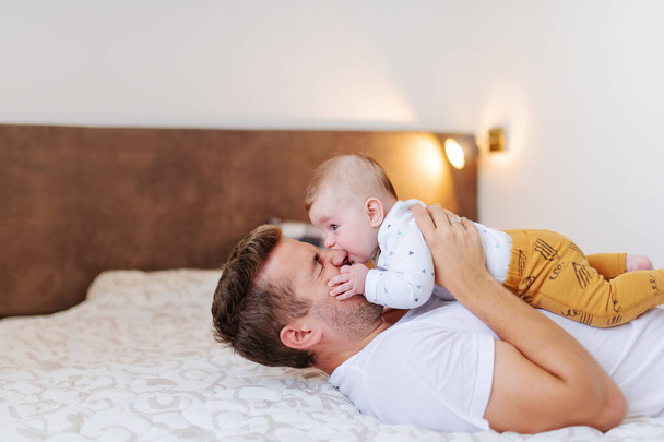 Smiling caucasian handsome dad lying on bed in bedroom and holding his loving adorable six months old son on his chest. Baby biting dad's nose. Unconditional love concept. - Photo, Image