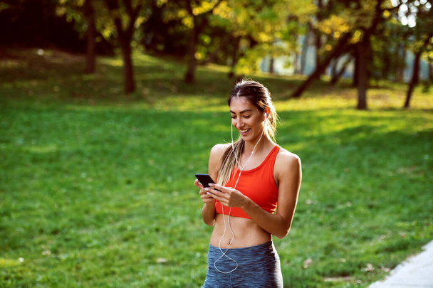 Attractive fit caucasian brunette in sportswear and with ponytail standing in nature, using smart phone and having earphones in ears. Sunny day in nature. - Photo, Image