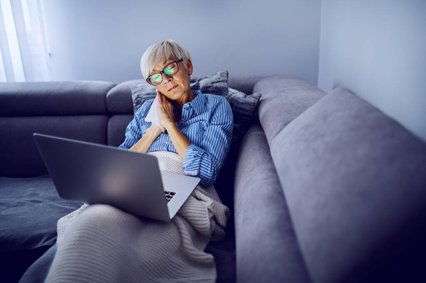 Charmante kaukasische blonde Seniorin mit Brille, die auf dem Sofa im Wohnzimmer sitzt, Laptop im Schoß hält und traurigen Film guckt. Sonntag Nachmittag zu Hause. - Foto, Bild