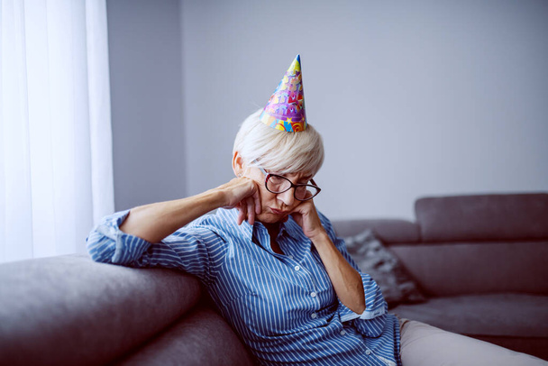 Θλιβερή καυκάσια ξανθιά ηλικιωμένη γυναίκα με καπέλο γενεθλίων στο κεφάλι κάθεται στον καναπέ στο σαλόνι. - Φωτογραφία, εικόνα