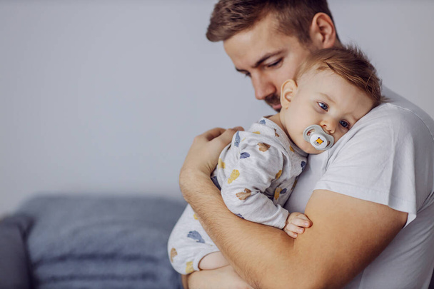 Sleepy adorable little boy with beautiful blue eyes having pacifier in his mouth and leaning on father's shoulder. Father is holding his beloved son and putting him to sleep. Nap time. - Photo, Image