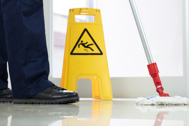 Een lage hoek close-up van een persoon het reinigen van de vloer met een dweil in de buurt van een gele waarschuwing natte vloer teken - Foto, afbeelding