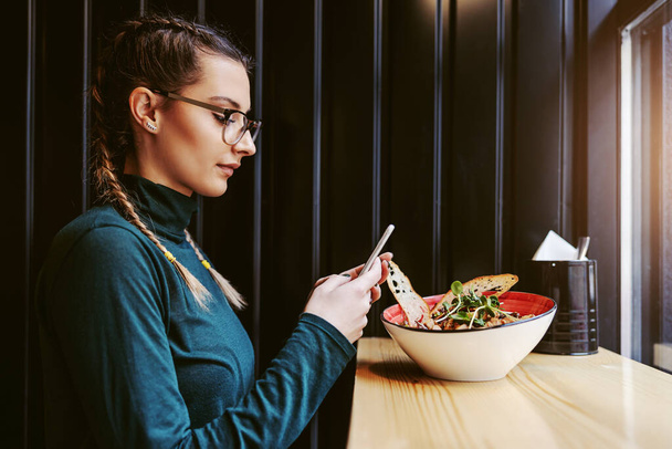 Vista lateral da mulher atraente com tranças sentadas no restaurante ao lado da janela e tirando fotos de seu almoço saudável
. - Foto, Imagem