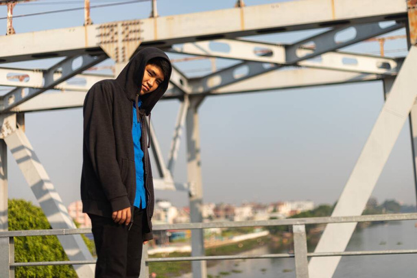 A low angle shot of a young Asian man standing on a bridge sadly looking at the camera - Фото, зображення