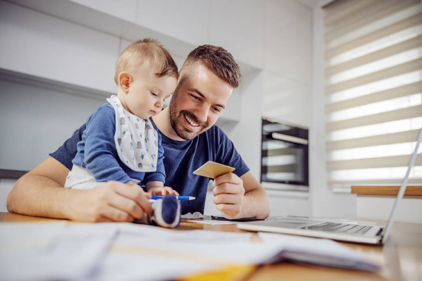 Young attractive bearded father sitting at dinning table with his beloved adorable son and paying bills online. His boy holding pen and trying to help him. - Photo, image