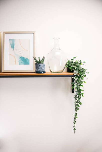 A vertical shot of a picture frame, house plants, and a transparent glass container on a white wall - Foto, Imagen