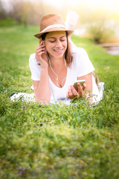 Mladá šťastná žena ležící v trávě a užívající si hudbu ze svého telefonu v přírodě. Usmívá se. Nošení klobouku. - Fotografie, Obrázek
