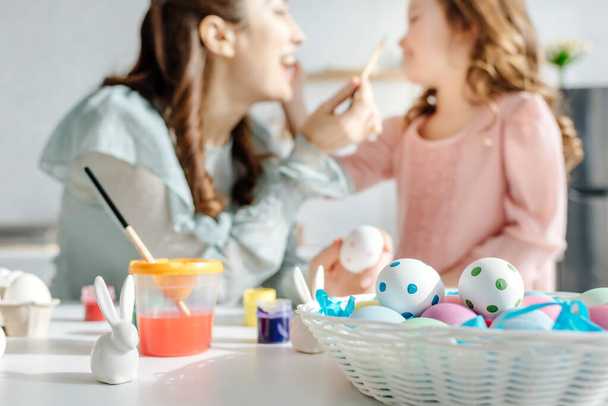 selective focus of painted easter eggs near decorative bunnies, happy mother and daughter  - Fotografie, Obrázek