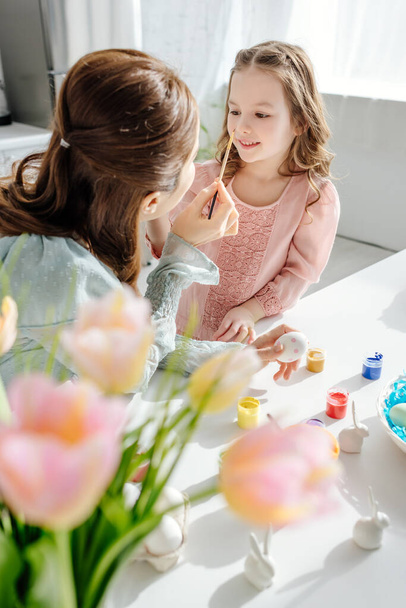 Selektiver Fokus des glücklichen Kindes auf die Mutter in der Nähe von Tulpen und dekorativen Hasen  - Foto, Bild