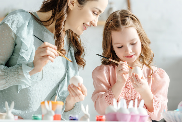 attractive mother and happy child painting easter eggs near decorative bunnies  - Photo, Image