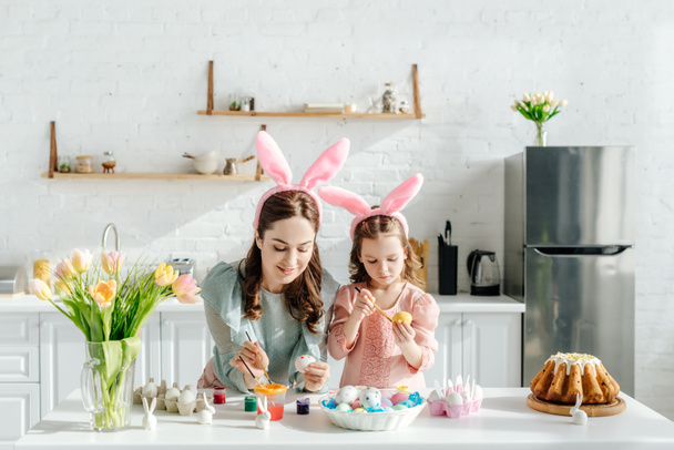 happy kid and attractive mother with bunny ears near chicken eggs, decorative rabbits, easter bread and tulips  - Zdjęcie, obraz