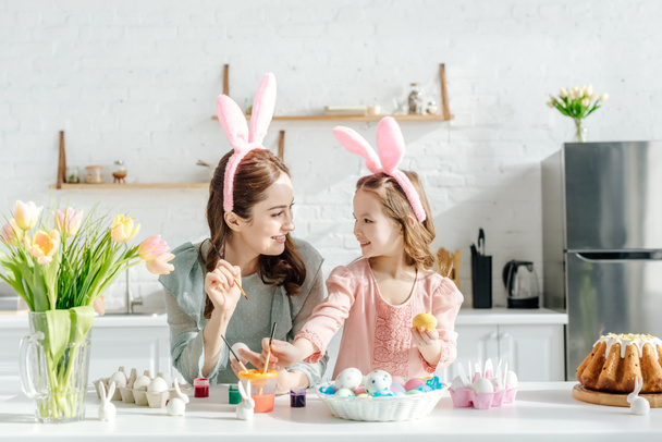 happy child and mother with bunny ears near chicken eggs, decorative rabbits, easter bread and tulips  - Photo, Image