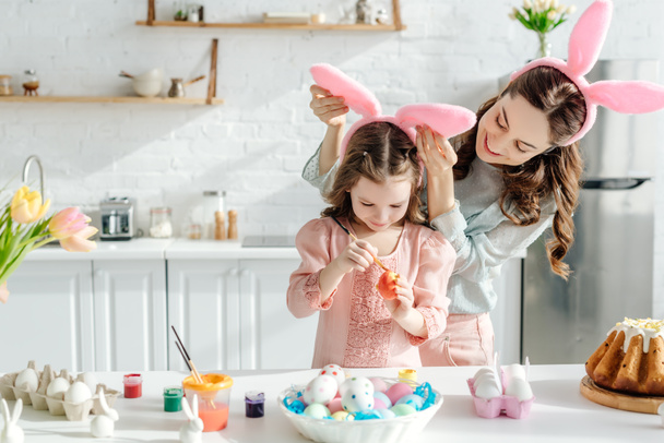 selective focus of happy mother touching bunny ears on daughter painting chicken egg  near easter bread  - Foto, afbeelding