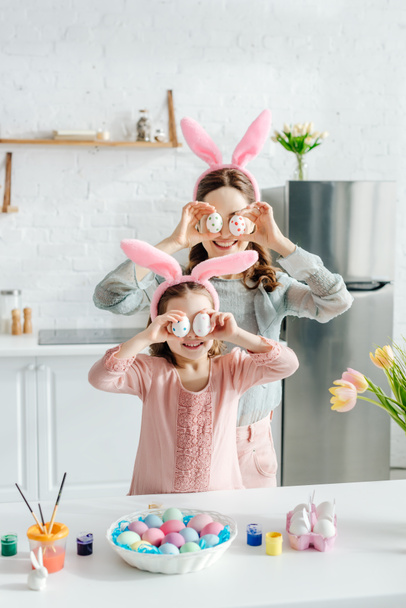 happy mother and daughter in bunny ears covering eyes with easter eggs near tulips  - Foto, Bild