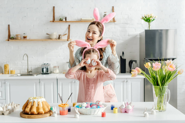feliz madre tocando conejito orejas en hija cubriendo ojos con huevos de pollo cerca de Pascua pan y tulipanes
  - Foto, imagen