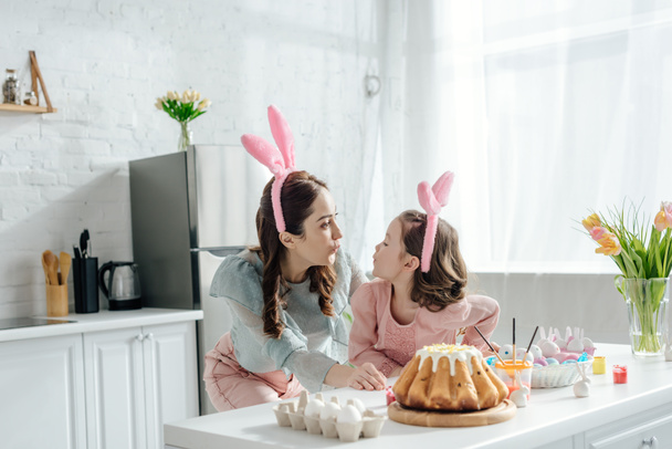 happy mother and daughter in bunny ears looking at each other near easter eggs - Fotografie, Obrázek