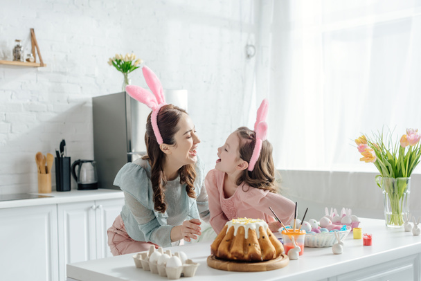 happy mother and daughter in bunny ears looking at each other near easter cake  - Fotografie, Obrázek