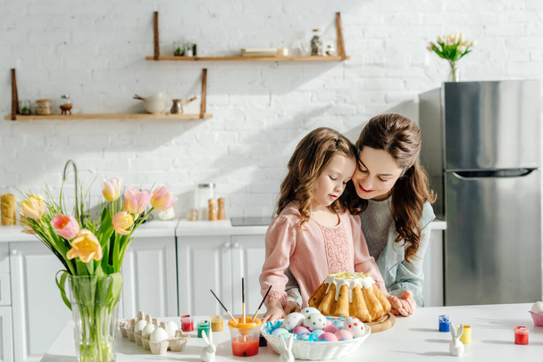 adorable child and happy mother near easter eggs, decorative rabbits, easter bread and tulips  - Foto, imagen