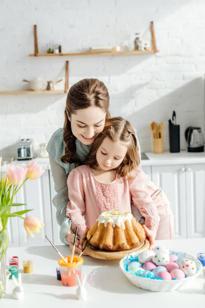 attractive mother and happy child near easter eggs, decorative rabbits, easter bread and tulips  - Foto, Imagem