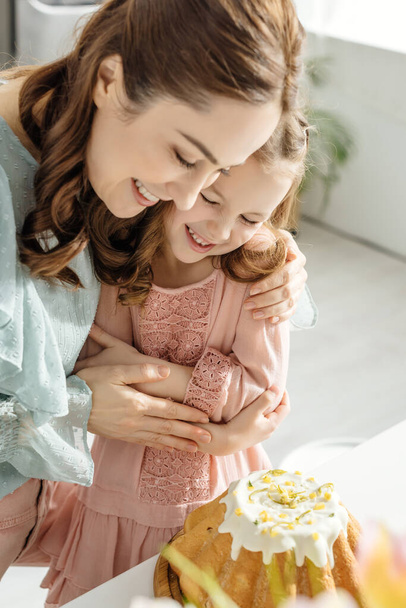 selective focus of happy woman hugging daughter while looking at easter cake  - Foto, afbeelding