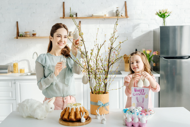 lindo niño sosteniendo huevos de Pascua decorativos cerca de sauce madre y conejos decorativos
  - Foto, imagen