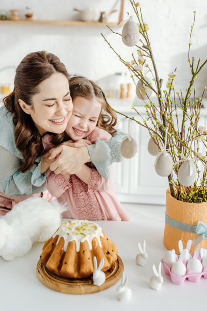 mãe feliz abraçando filha perto de vaso de flores willowin, coelhos decorativos e bolo de Páscoa
  - Foto, Imagem