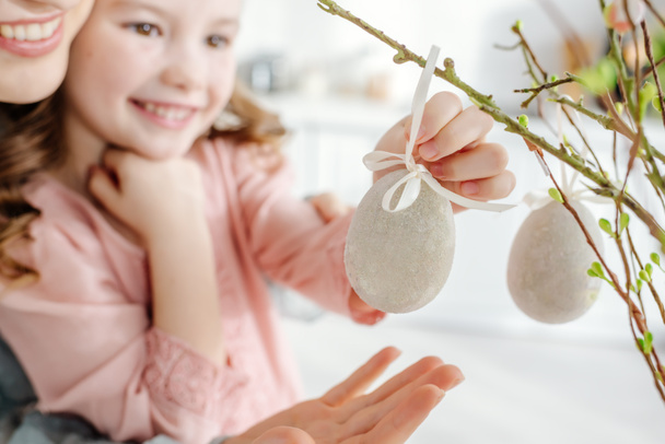 szelektív fókusz boldog gyerek megérinti dekoratív húsvéti tojás fűzfa ágon közel mosolygós anya  - Fotó, kép